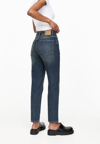 Jean taille haute, jambes droites, en denim bleu foncé. Présente des coutures en cuivre, un design de poche patch et une légère décoloration sur le tissu.
