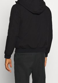 Černá kapucí mikina se zipem, žebrovanými manžetami a pasem; hladká textura látky, na levém rameni se nachází logo.