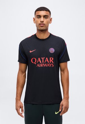 Jeune homme debout portant un maillot de football noir du Paris Saint-Germain avec le logo rouge de Qatar Airways à l'avant et le symbole Nike.