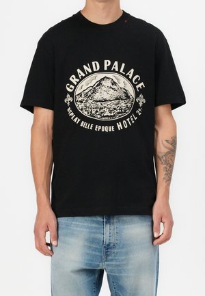 Homme portant un t-shirt noir avec un graphique de montagne et de ville et le texte "Grand Palace Replay Belle Epoque Hotel 29", associé à un jean bleu clair.