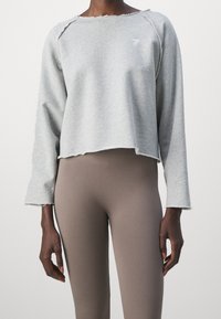 Sweatshirt gris court avec ourlet brut et manches longues, associé à des leggings bruns côtelés. Présente un logo subtil sur la poitrine.
