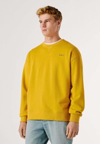 Jeune homme aux cheveux blonds courts portant un sweat-shirt jaune moutarde et un jean bleu clair, debout contre un fond uni.