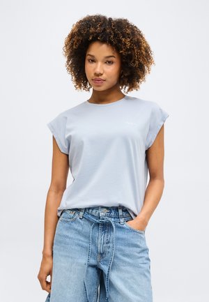 Νεαρή γυναίκα με σγουρά μαλλιά, φοράει ανοιχτό μπλε αμάνικη μπλούζα και μπλε τζιν ψηλόμεσο με δεμένη ζώνη, χέρι στην τσέπη.