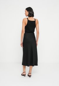 Robe noire sans manches avec un corsage ajusté et une jupe fluide, présentant une texture lisse. Portée avec des talons bordeaux.