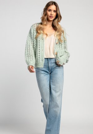 Jasnozielony dziergany cardigan o luźnym, otwartym splocie, z drewnianymi guzikami, zestawiony z kremową bluzką i jasnoniebieskimi dżinsami.