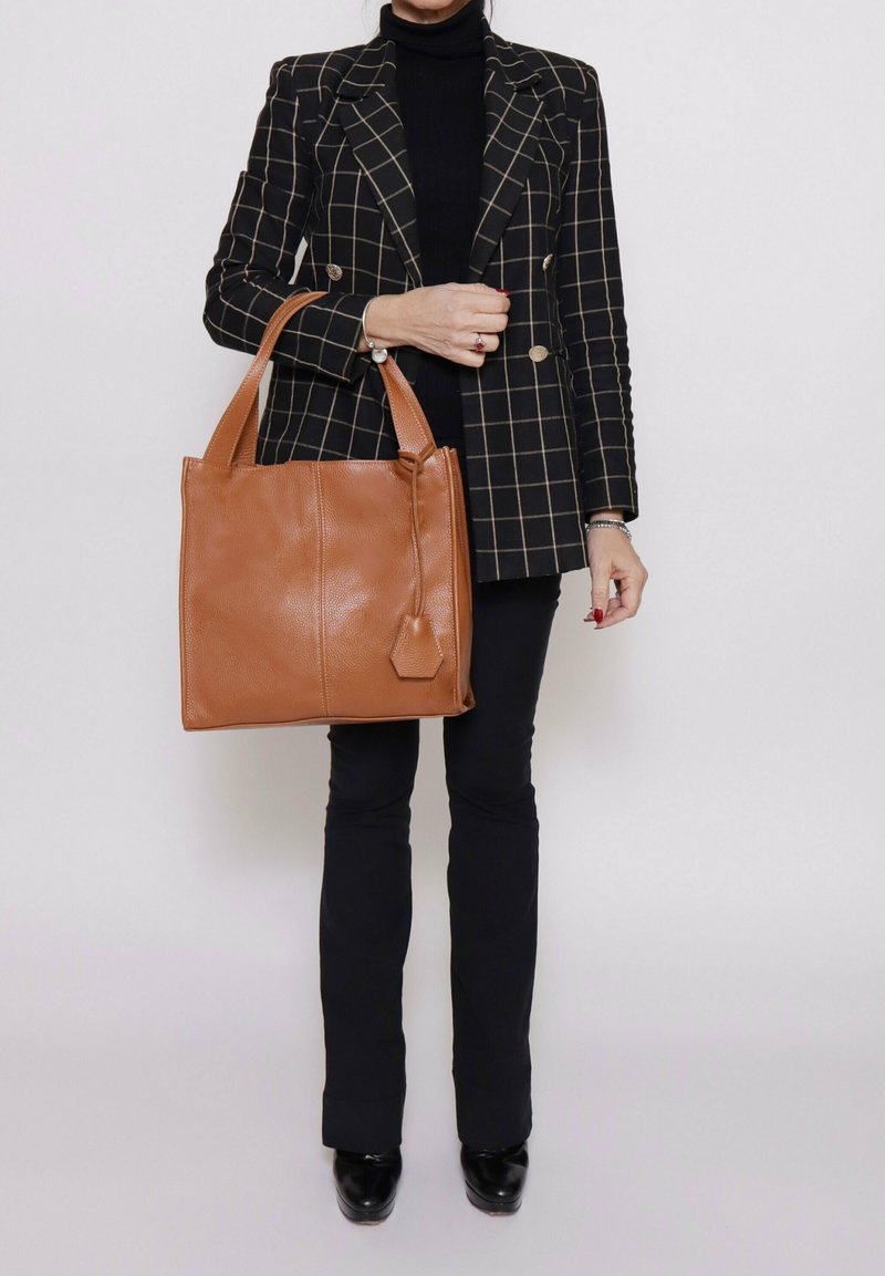 Borsa tote in pelle marrone con una finitura testurizzata, due manici robusti e un'etichetta decorativa, abbinata a un blazer a quadri neri e pantaloni neri.