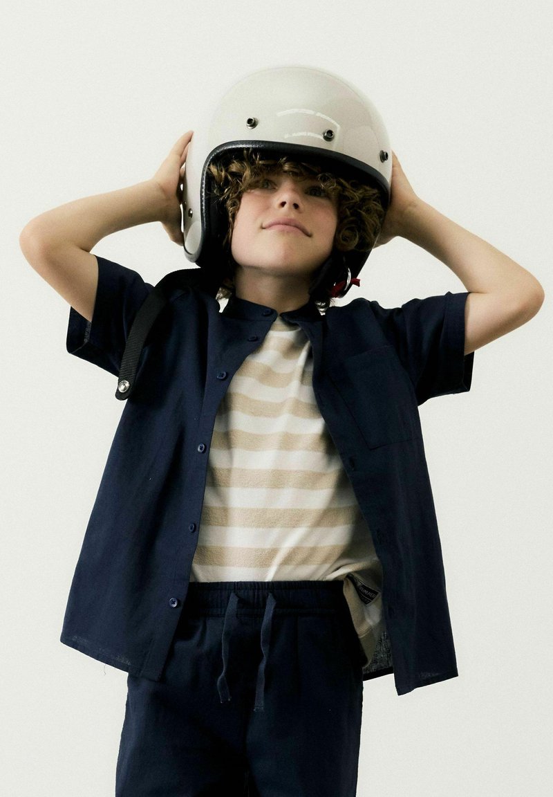 Niño vistiendo una camisa de botones azul marino sobre una camiseta a rayas beige y blanca y pantalones azul marino a juego, sosteniendo un casco blanco brillante.