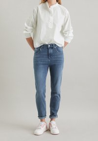 Valkoinen puuvillapaita pystykauluksella, yhdistettynä slim fit -sinisiin farkkuihin. Valkoiset lenkkarit, joissa on vaaleanpunaisia vivahteita. Rentoa, modernia tyyliä.