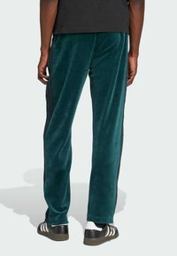Persona vistiendo pantalones de chándal de terciopelo color teal con rayas laterales negras y zapatillas en blanco y negro, mostrada desde atrás contra un fondo liso.