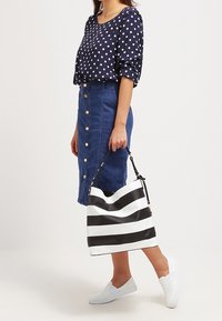 Femme portant un chemisier bleu marine à pois, une jupe en jean boutonnée, des chaussures blanches sans lacets, tenant un sac à main rayé noir et blanc à côté d'elle.