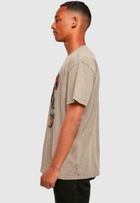 Oversized, light brown cotton t-shirt with a crew neck, featuring a graphic print on the left side. Short sleeves, relaxed fit, soft texture.