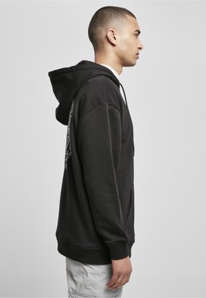 Junger Mann im Profil, der einen schwarzen Hoodie mit bedrucktem Design auf der Rückseite und hellfarbene Hosen vor einem einfachen Hintergrund trägt.