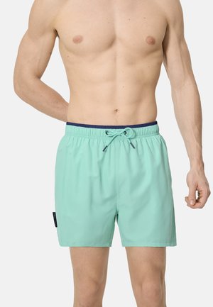 Hombre sin camisa con pantalones cortos de baño de color turquesa claro, con cintura elástica azul marino y cordón, de pie contra un fondo liso y claro.