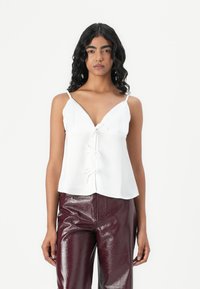 Top bianco senza maniche con scollatura a V, dotato di chiusura con bottoni e lacci in tessuto. Abbinato a pantaloni a vita alta bordeaux lucidi.