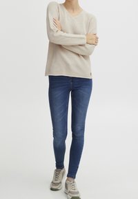 Pull beige tricoté avec un col en V et des manches longues, associé à un jean skinny bleu et des baskets grises avec des accents verts.