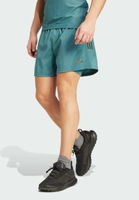 Man in teal sportshorts, grijze enkelkousen en zwarte hardloopschoenen die tegen een lichte achtergrond staat.