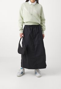 Jasnozielona bluza z dużym logo Nike, czarna długa spódnica maxi, teksturowana czarna torebka i białe trampki z niebieskimi akcentami.