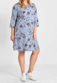 Vestido floral azul con mangas tres cuartos, que presenta una variedad de patrones de flores rosas y moradas sobre una tela suave. Usado con zapatillas blancas.