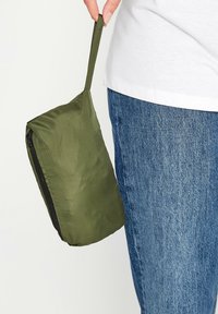 Pochette leggera di colore verde oliva con chiusura a zip e manico in tessuto, attaccata al fianco di una persona. Jeans di denim visibili in basso.