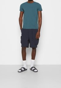 Tyrkysové bavlněné tričko s kulatým výstřihem, doplněné o námořnické cargo šortky s bočními kapsami. Oblečeno s bílými ponožkami do poloviny lýtek a černými sandály.