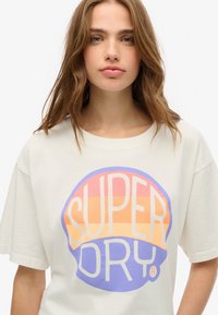 Camiseta color crema con un gran gráfico circular que presenta el texto "SUPER DRY" en rayas moradas y naranjas. Ajuste casual con mangas cortas.