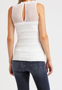 Top bianco senza maniche con schiena trasparente, strisce orizzontali testurizzate e un piccolo dettaglio a chiave sul collo. Abbinato a jeans scuri.