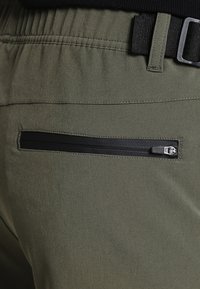Olijfgroene broek met een zwarte ritszak, elastische tailleband en gestructureerde stof. Zichtbare riemlussen en gladde naden.