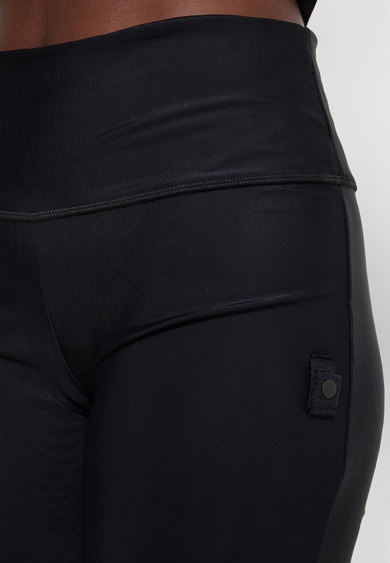 Schwarze, hochtaillierte Leggings aus glattem, elastischem Material, mit einem strukturierten Patch und einem Druckknopf an der Seite.