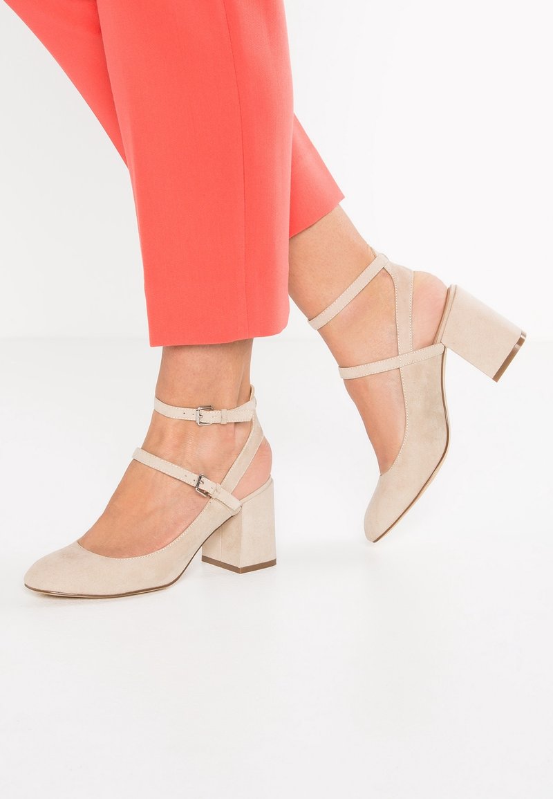 Chaussures à talons blocs en suède beige avec double bride de cheville et boucles en argent, portées avec un pantalon corail sur fond blanc.