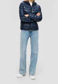 Granatowa ocieplana kurtka z kapturem, zapinana na zamek z przodu, z poziomym pikowaniem i dwoma bocznymi kieszeniami, w połączeniu z jasnoniebieskimi rozszerzanymi dżinsami i białymi trampkami.