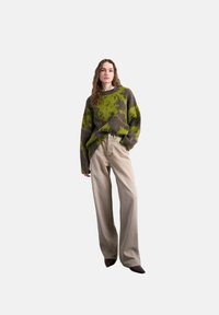 Femme portant un pull à motifs verts et bruns, un pantalon beige à jambes larges, et des chaussures pointues foncées, debout avec une main dans la poche.