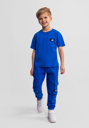 Młody chłopiec idący do przodu, ubrany w jasnoblękitną koszulkę i pasujące spodnie typu cargo oraz białe trampki, na tle jednolitego szarego tła.