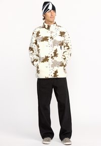 Veste en polaire crème avec des taches brunes et grises, fermeture éclair sur le devant, col montant et poches latérales. Portée avec un pantalon noir à jambes larges et des chaussures à carreaux.