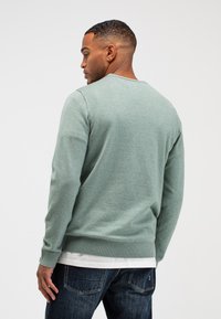 Grüner gestrickter Pullover mit Rundhalsausschnitt, gerippten Bündchen und Saum, mit einer strukturierten Oberfläche. Kombiniert mit dunkler Jeans.