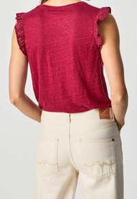 Žena v bezrukávovém červeném topu s volánkovými průramky a vysokými off-white džíny s přišitými zadními kapsami a koženou etiketou.