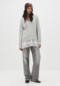 Szary sweter dzianinowy z dekoltem w serek, nałożony na biały top z koronkowym wykończeniem. W zestawie z wyblakłymi szarymi spodniami o szerokich nogawkach i czarnymi butami. Brązowa chustka na głowie.