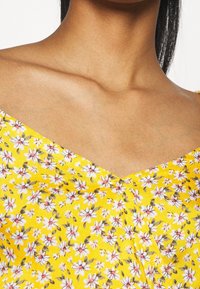 Gul blommig klänning med v-ringning, prydd med små vita och rosa blommor på en texturerad tyg, som framhäver ett somrigt blommönster.