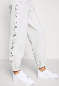 Ljust grå joggers med "NIKE SPORTSWEAR" tryckt i svart längs sidan, med en avslutad kant. Matchas med vita Nike Air Force 1-sneakers.