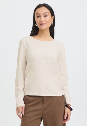 Kvinde med langt mørkt hår iført en beige langærmet top og brune bukser, står foran en ensfarvet, lys baggrund og kigger fremad.
