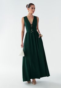 Lange, donkergroene jurk met een diep uitgesneden halslijn, een getailleerde taille en een A-lijn silhouet. Voorzien van een strikdetail en gouden accenten.