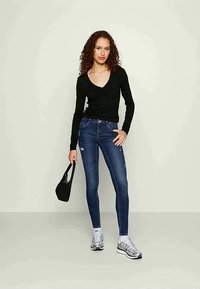 Top preto de manga longa combinado com jeans skinny azul desgastados. O modelo segura uma pequena bolsa preta, usando ténis prateados com detalhes brancos.