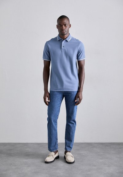 Cămașă polo de culoare albastru deschis, cu guler și accente albe, asortată cu blugi bleumarin și mocasini bej. Modelul stă pe un fundal neutru.
