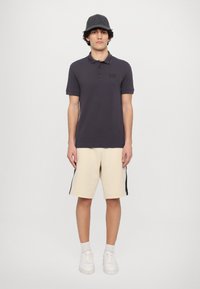 Polo oscuro de color gris con mangas cortas y detalle de logo. Pantalones cortos beige con franjas laterales negras. El modelo lleva zapatillas blancas y una gorra gris.