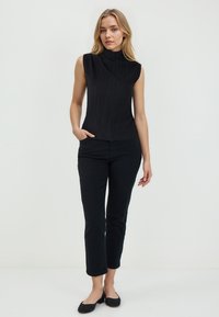 Zwarte mouwloze top met een hoge kraag en ribbeltextuur, gecombineerd met zwarte broek tot de enkels en zwarte flats. Over het algemeen een minimalistisch ontwerp.