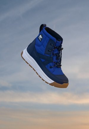 Botte d’hiver bleue et noire avec semelle blanche et bande de roulement marron, avec lacets et écusson logo, présentée devant un ciel nuageux.