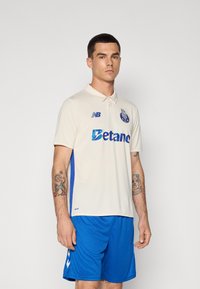 Polo crème avec des panneaux latéraux bleus, arborant le logo New Balance et le mot "Betano" en texte iridescent. Porté avec un short bleu.