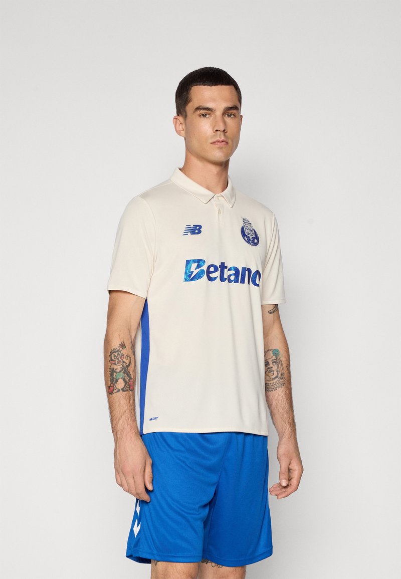Polo crème avec des panneaux latéraux bleus, arborant le logo New Balance et le mot "Betano" en texte iridescent. Porté avec un short bleu.