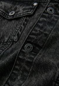 Tessuto in denim nero con una superficie strutturata, dotato di bottoni a pressione in metallo argentato e dettagli di cucitura a contrasto su un pannello della giacca.