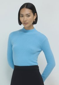Hellblauer, strukturierten Strickpullover mit hohem Kragen, kombiniert mit einem dunklen Rock. Der Stoff wirkt weich und hat eine lässige Passform.
