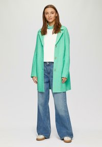 Abrigo de mezcla de lana verde con cuello, tres botones y corte relajado, combinado con vaqueros de pierna ancha azul claro y zapatillas beige.